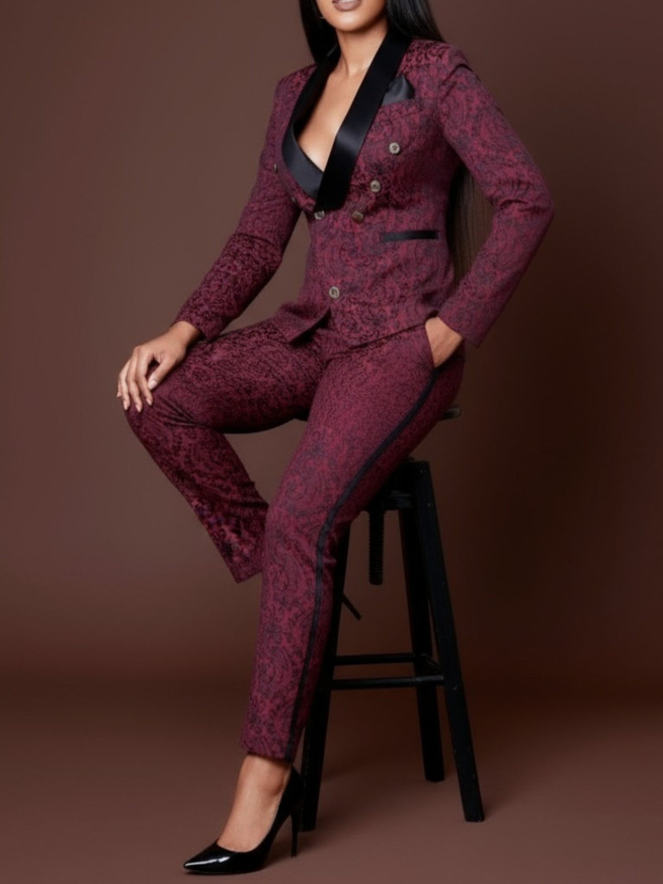 Woman wearing a maroon patterned suit with black accents sitting on a stool against a brown background
