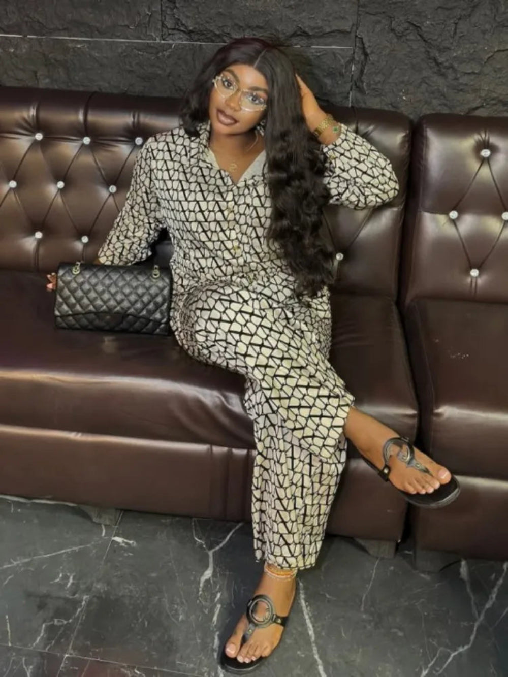 A woman in patterned outfit sitting on a brown leather couch with a dark marble wall background