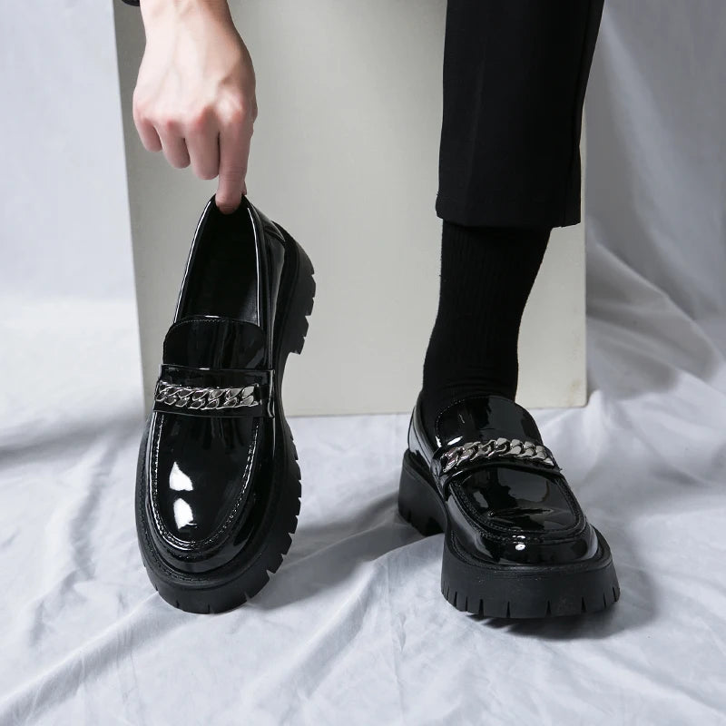 Black glossy loafers with chain details worn by a person on a white surface.