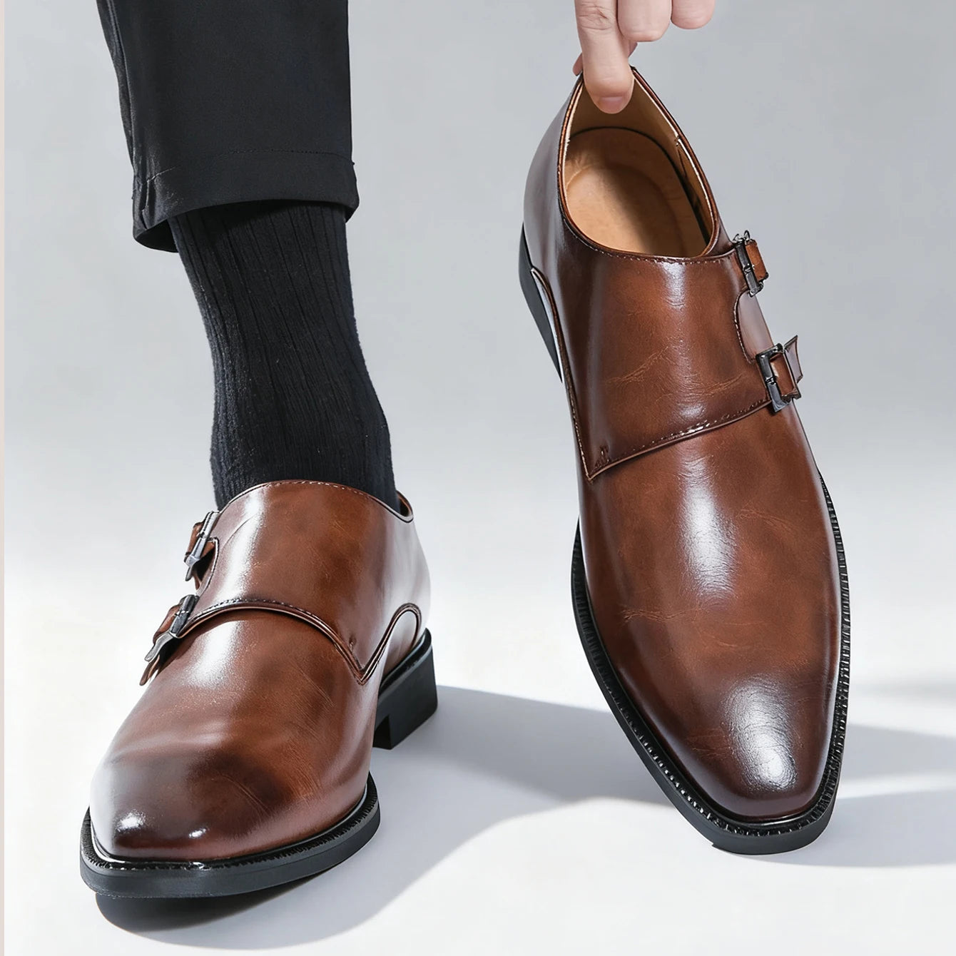 Brown leather dress shoes with buckles held by a person wearing black socks and pants.