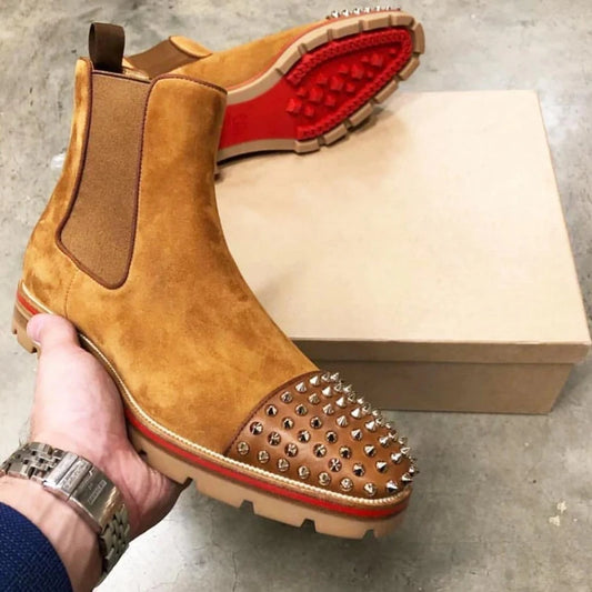A brown suede boot with a studded design is held by a hand, with a red sole and beige box in the background.