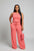 A woman in a coral jumpsuit is standing in front of a mirror with draped curtains.