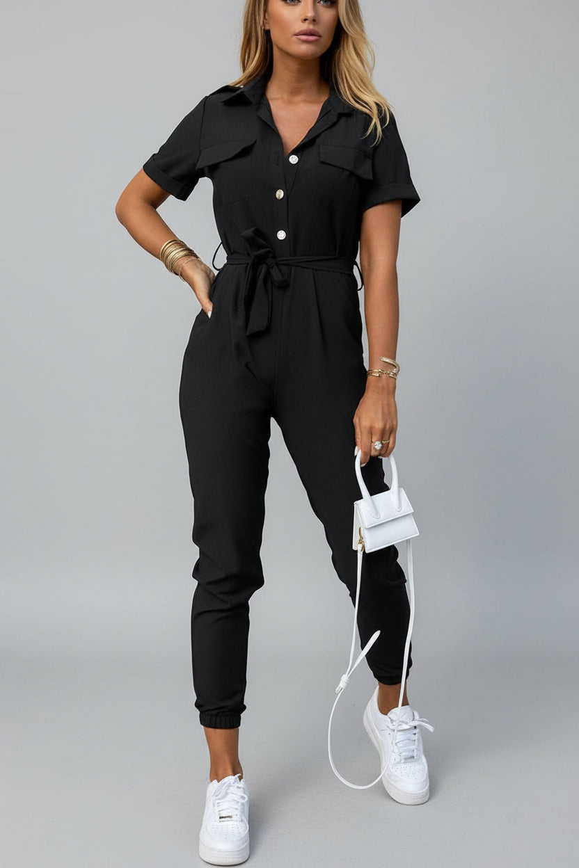 Woman in black jumpsuit, white sneakers and small white handbag against beige wall.