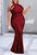 A woman in a burgundy lace dress with cut-out shoulders stands on a sidewalk.