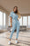 A woman in a light blue jumpsuit stands before a beige wall.