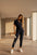Woman in navy jumpsuit with white sneakers and small white handbag, against a beige wall.