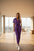 Woman wearing a purple jumpsuit with a white handbag against a beige wall.