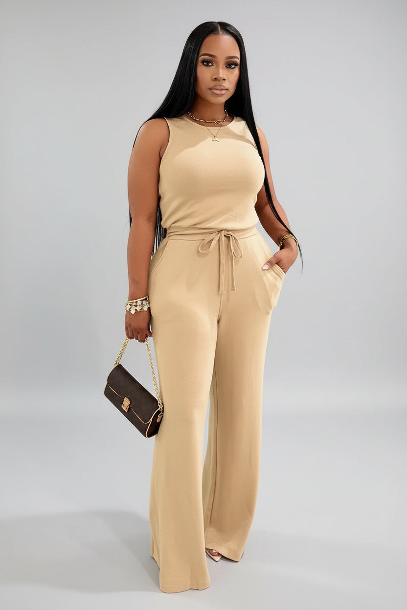 A woman wearing a beige jumpsuit is standing in front of draped curtains.