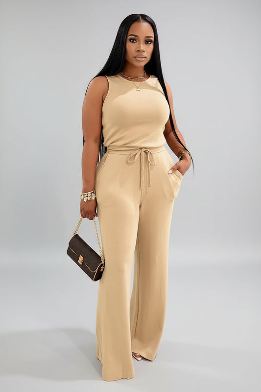 A woman wearing a beige jumpsuit is standing in front of draped curtains.