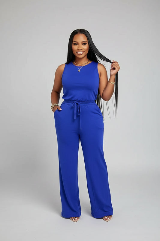 A woman wearing a blue jumpsuit is standing in front of a mirror with curtains.