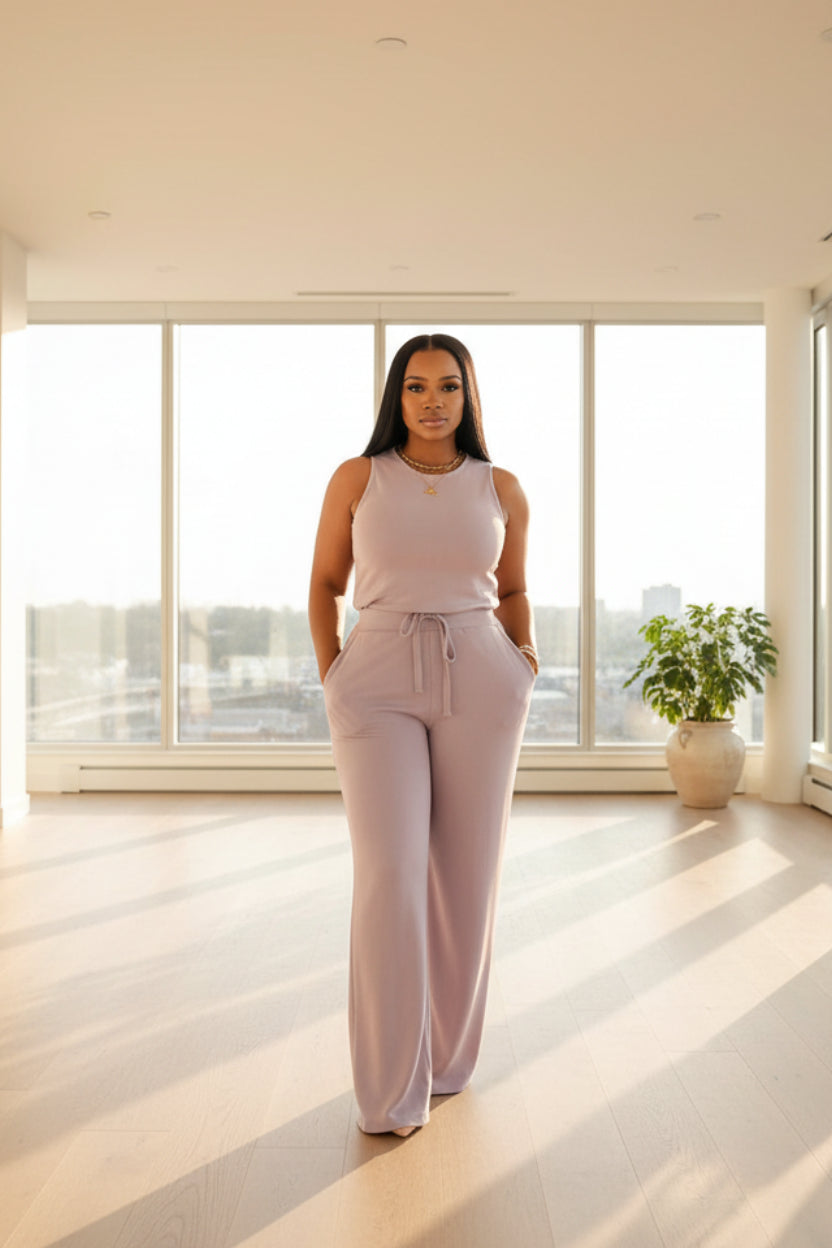 A woman wearing a light purple jumpsuit is standing in a room with curtains.