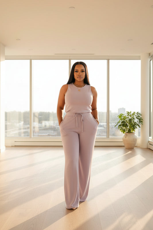 A woman wearing a light purple jumpsuit is standing in a room with curtains.