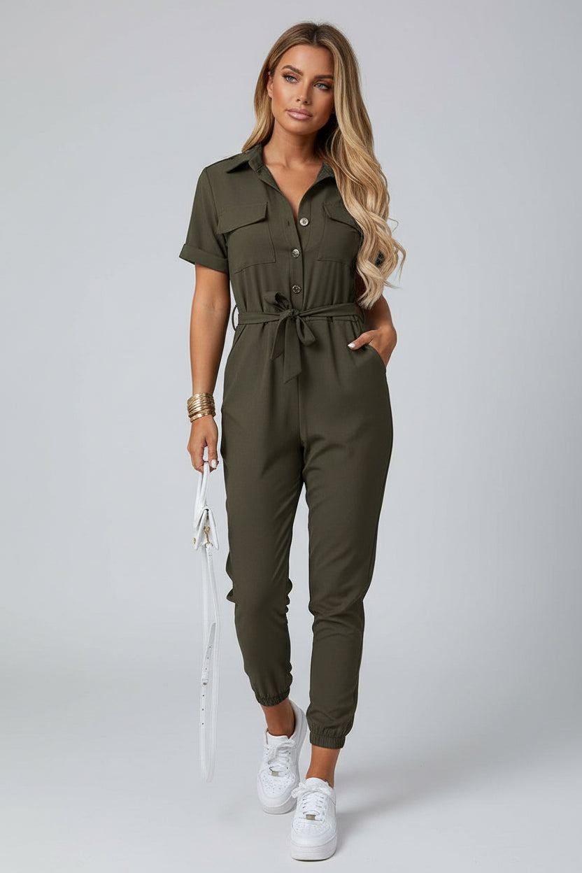 Woman wearing an olive green jumpsuit with a white handbag against a stone wall.
