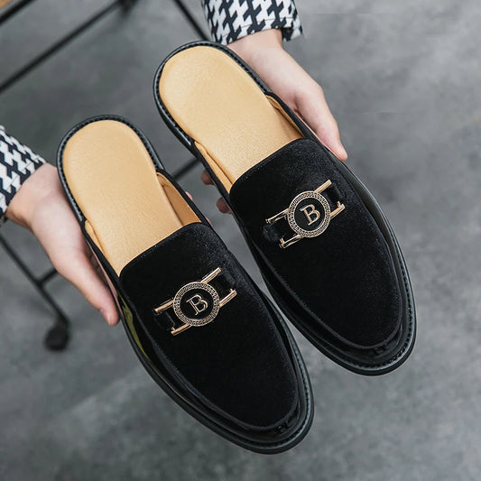 Black velvet loafers with gold 'B' emblem held by a person against a gray background