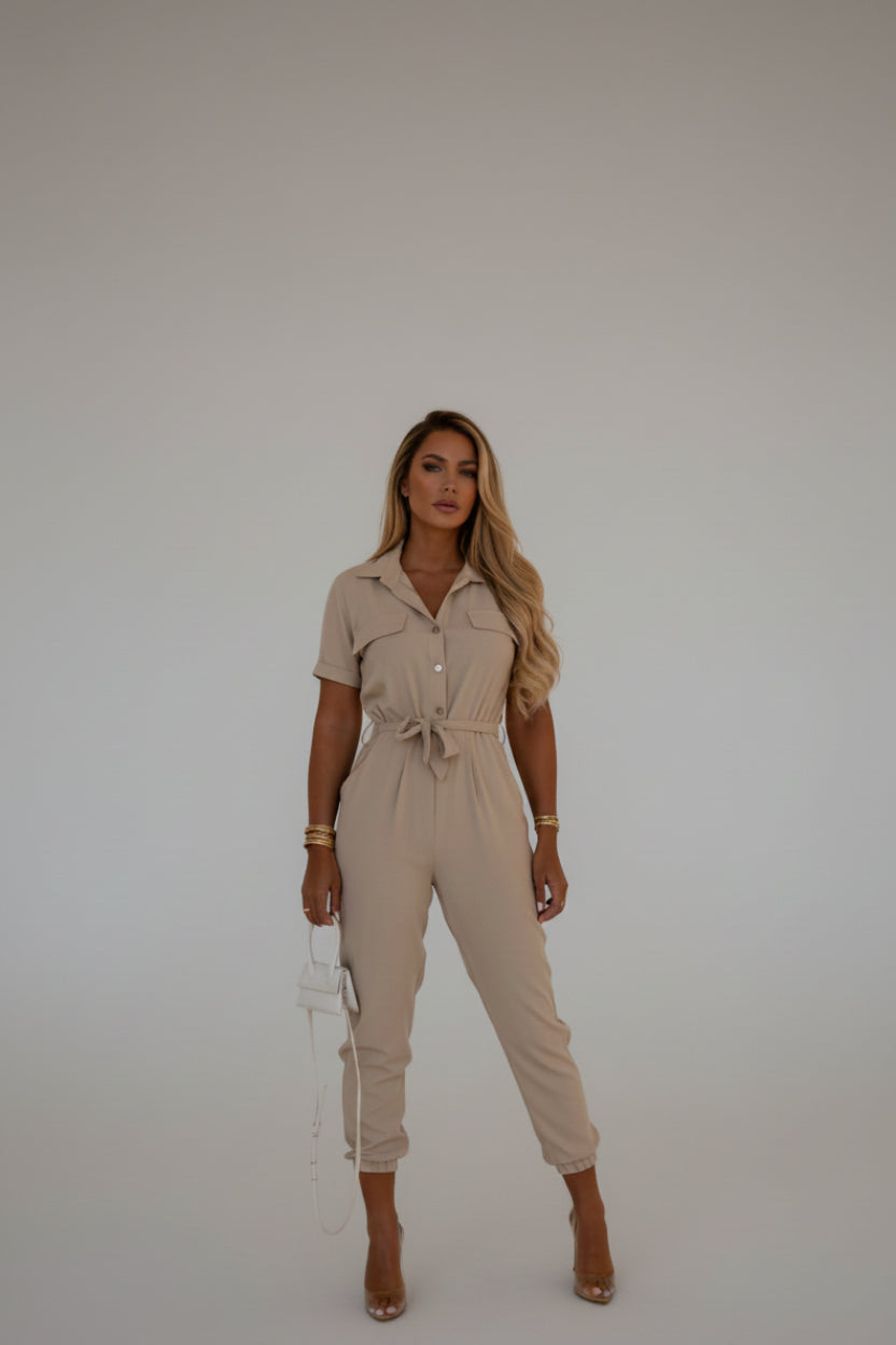 A woman wearing a beige jumpsuit is standing against a beige wall.