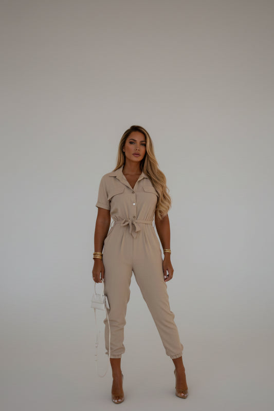 A woman wearing a beige jumpsuit is standing against a beige wall.